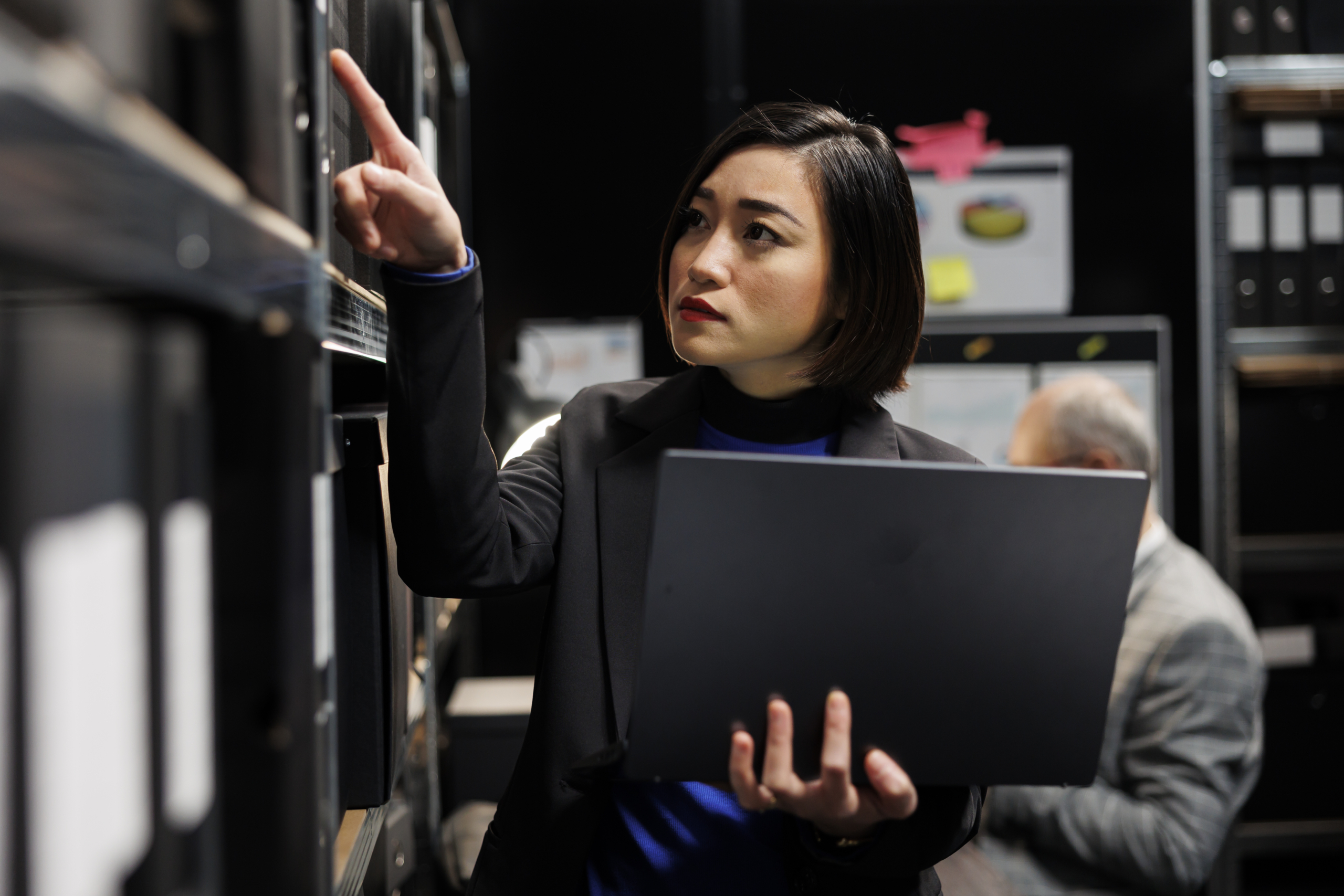 A woman holding a laptop points toward a shelf of files in an office, appearing focused while reviewing records. A woman holding a laptop points toward a shelf of files in an office, appearing focused while reviewing records.
