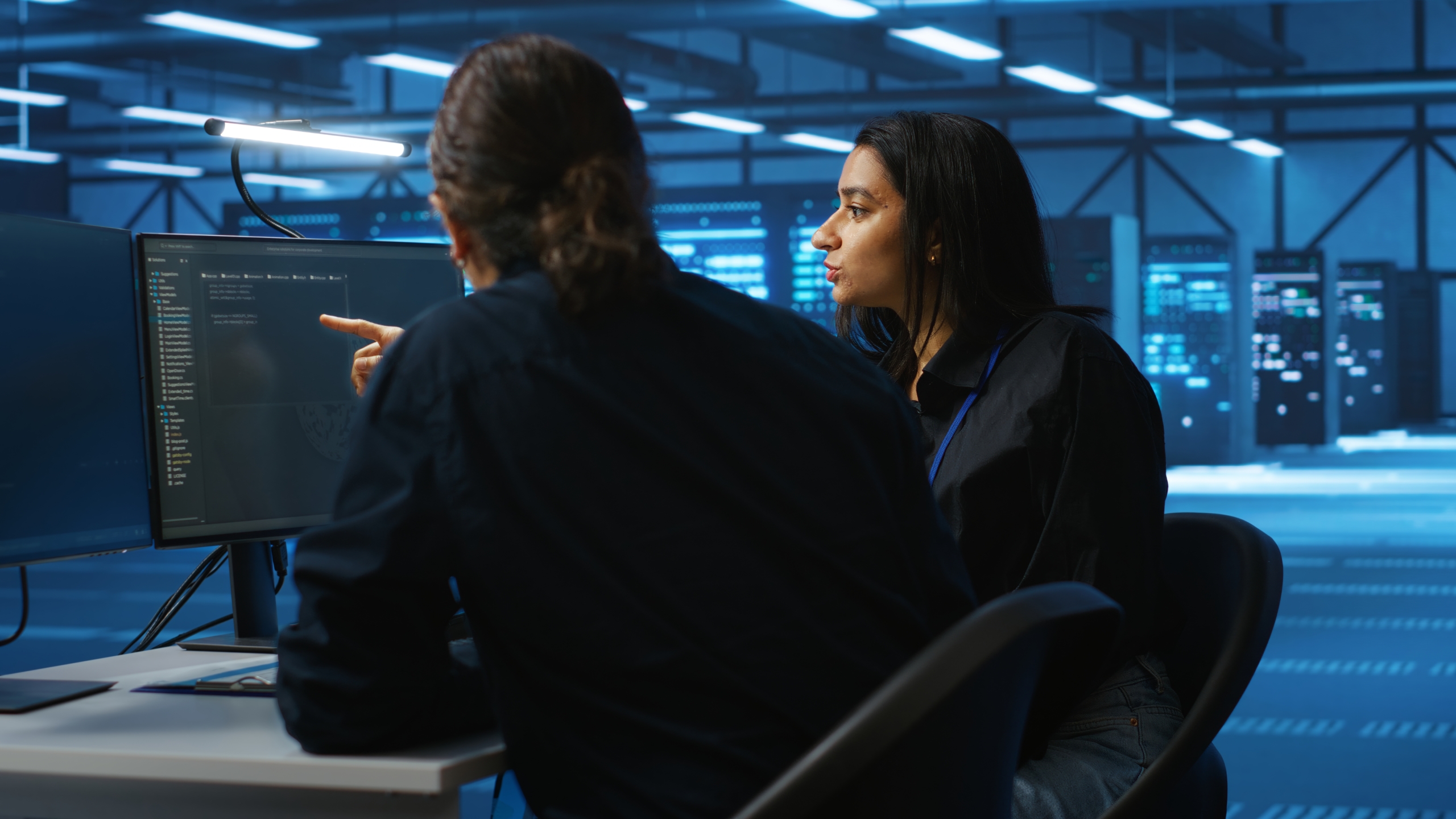 Two IT professionals sit at a desk in a server room having a discussion in front of a computer monitor. Two IT professionals sit at a desk in a server room having a discussion in front of a computer monitor.