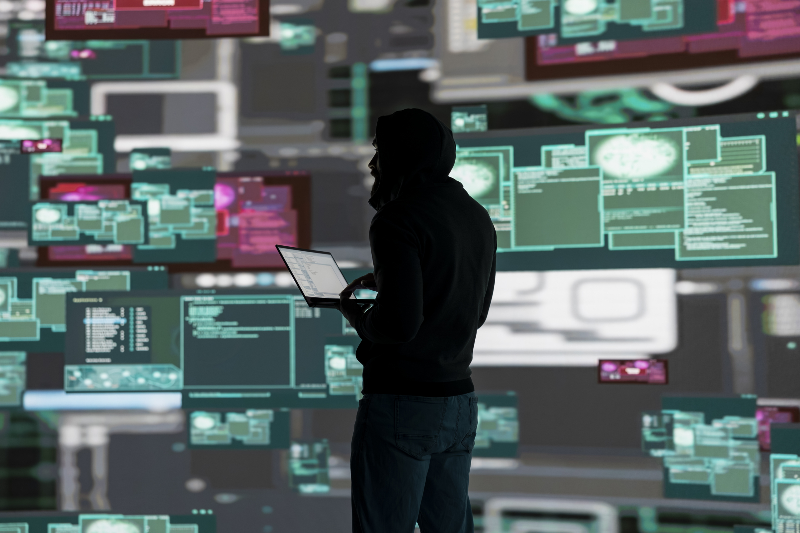 A person wearing a hooded sweatshirt works on a laptop surrounded by floating digital screens displaying code and data in a cybersecurity environment. A person wearing a hooded sweatshirt works on a laptop surrounded by floating digital screens displaying code and data in a cybersecurity environment.