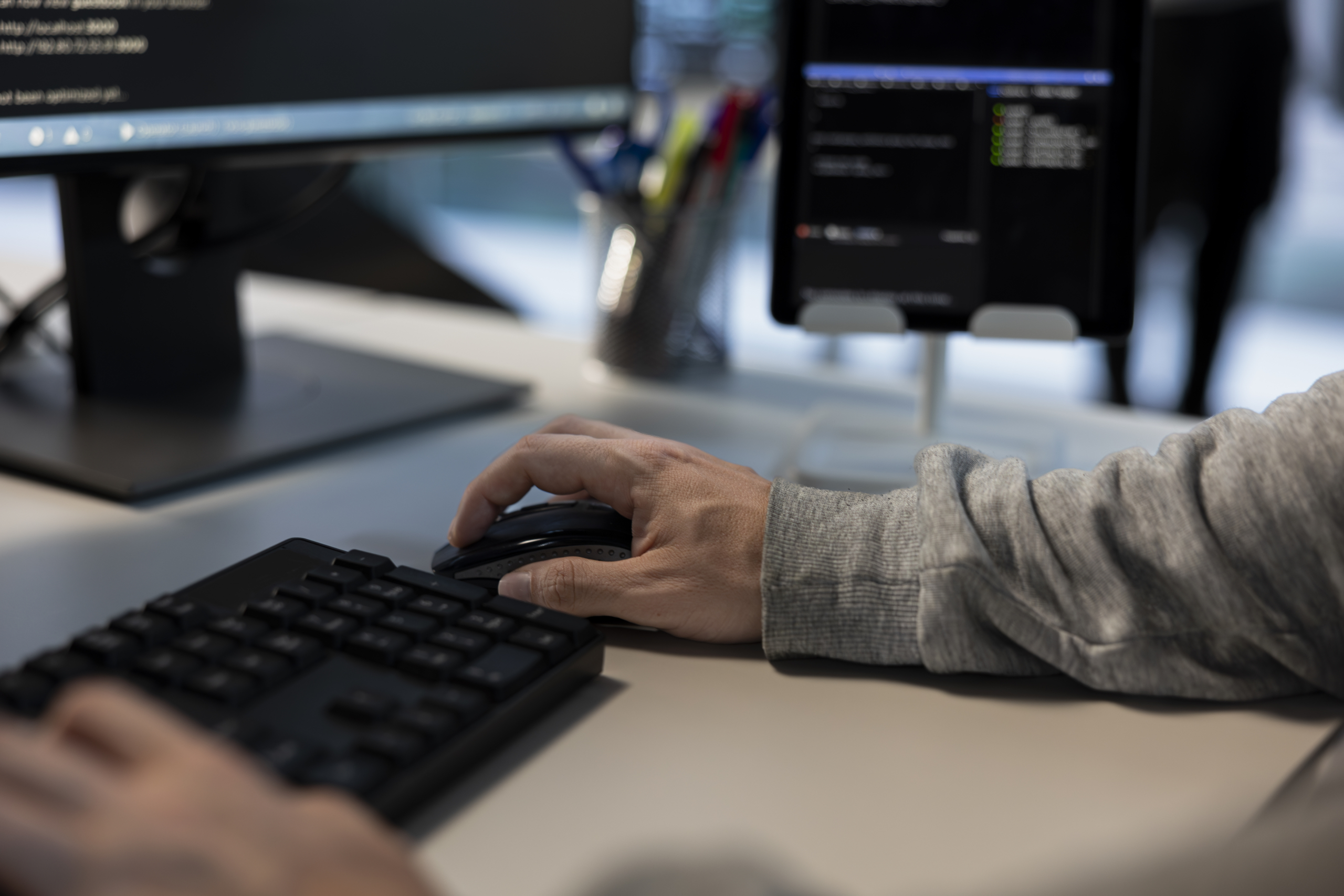 Hand on computer mouse beside keyboard and multiple monitors.

